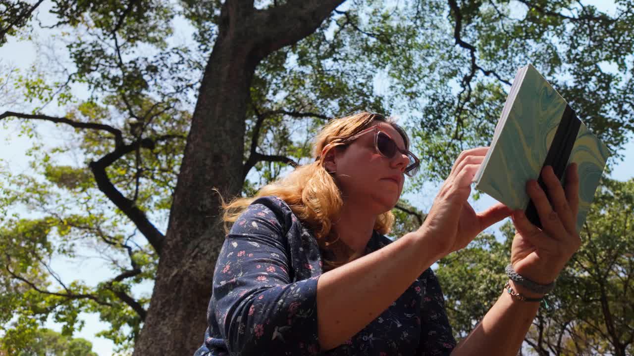 Low-angle shot of woman using modern sunglasses with anti-blue light and UV filters to protect her eyes while enjoying a book in the bright, sun-dappled shade of park, visual wellness