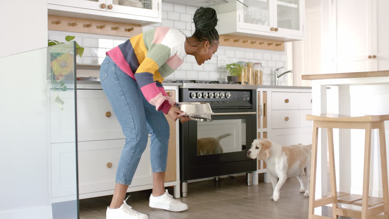African American young woman feeding dog in kitchen, at home