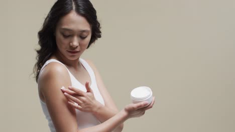 asian woman with dark hair applying cream on her skin on beige background, copy space, slow motion