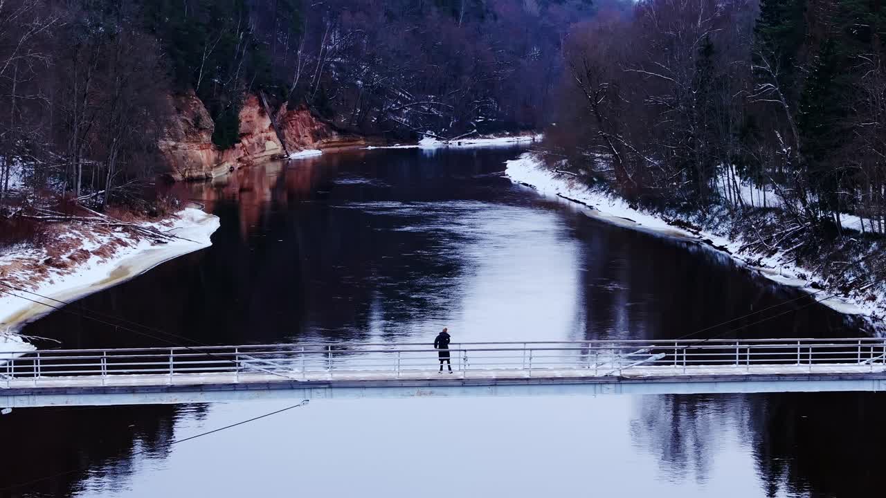 Frozen Gauja flows beneath woman dancing in solitude on pedestrian bridge