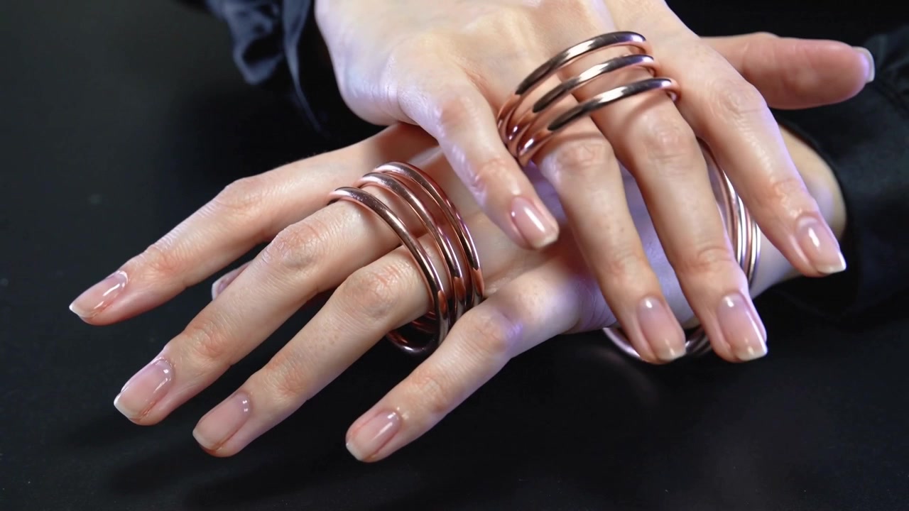 Apertura di un tavolo che rivela mani femminili con maniche scure che girano e mostrano anelli a spirale di rame e un braccialetto.