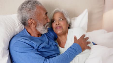 Happy-senior-biracial-couple-lying-on-bed,-embracing-and-talking,-unaltered,-in-slow-motion