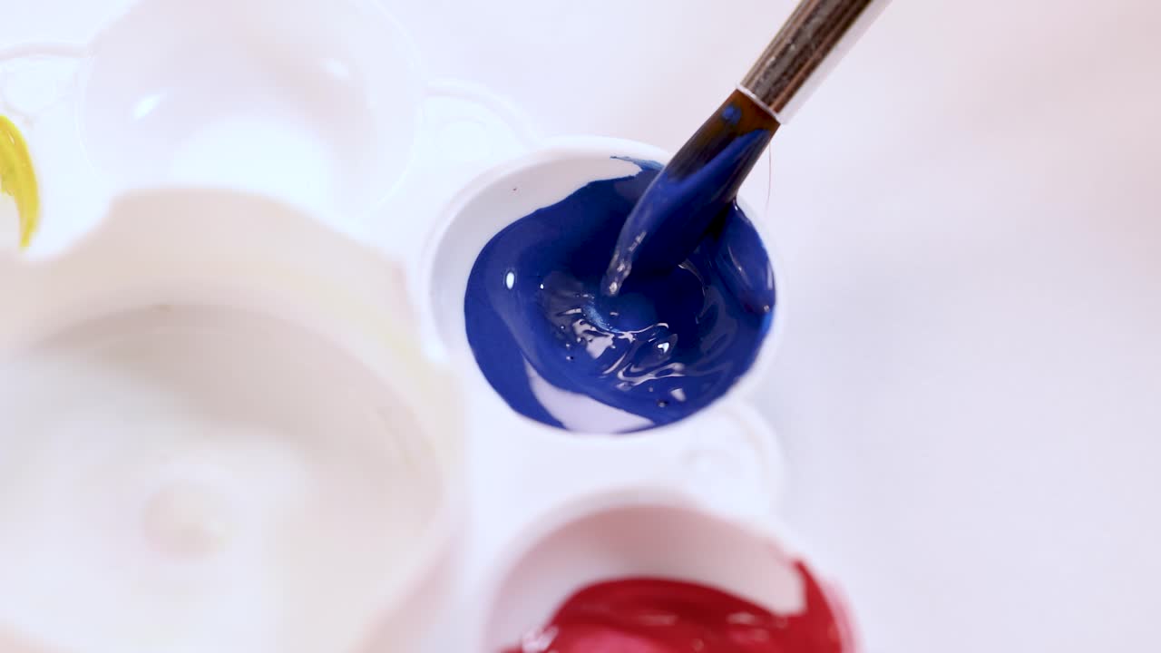 Paintbrush dips into blue paint in white palette, bright lighting, overhead close-up, minimal movement