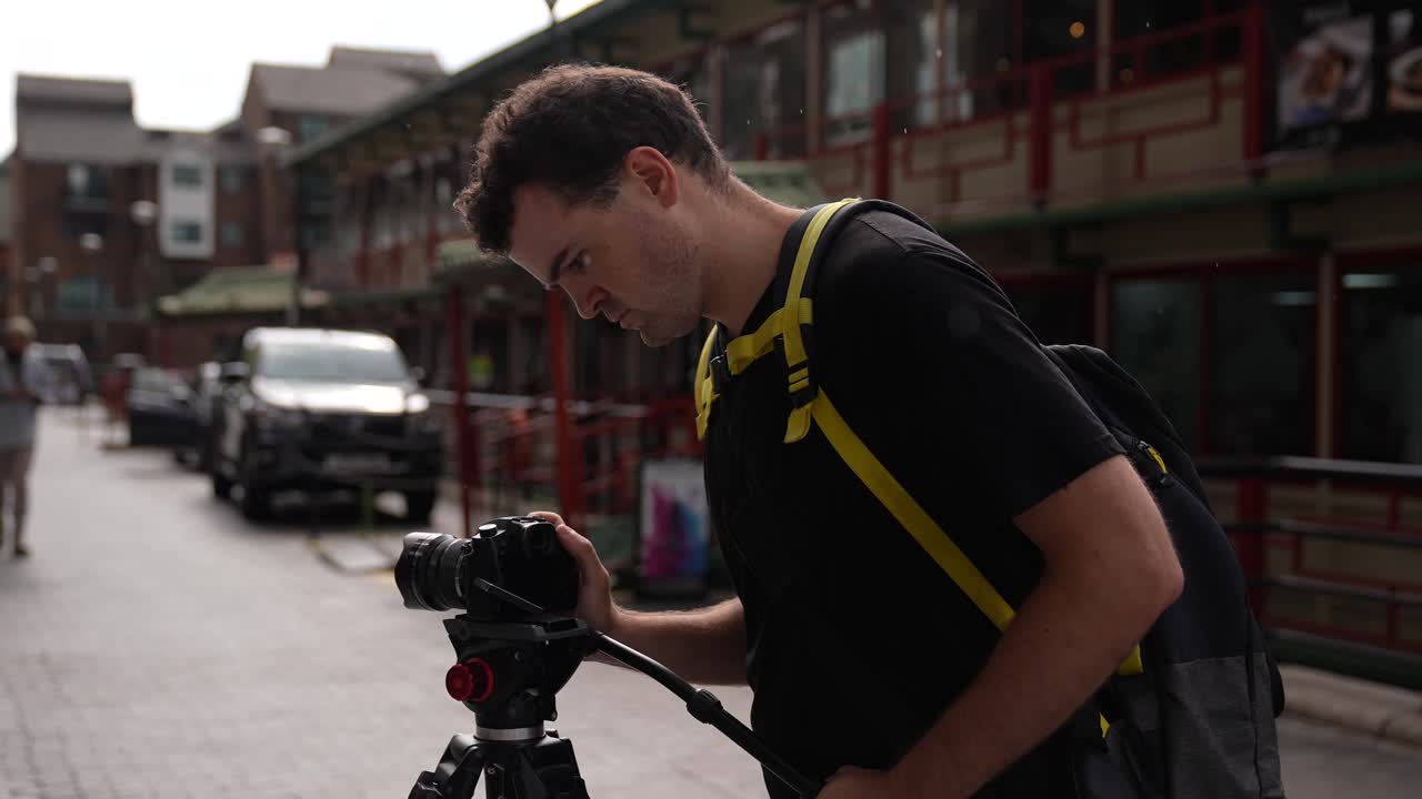 Young caucasian man is standing with his camera on a tripod and adjusting the settings for his shot