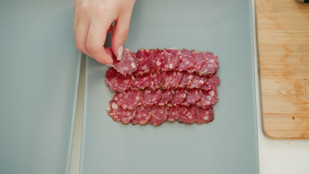 Hands placing rows of thin cured sausage slices on sleek tray, showcasing rich red marbling, precise alignment, gourmet appetizer styling, modern plating technique for elegant food presentation