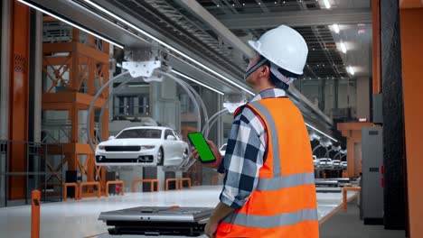 vista trasera de un ingeniero masculino asiático con casco de seguridad trabajando en un teléfono inteligente de pantalla verde y mirando a su alrededor mientras está de pie en una fábrica de automóviles ev