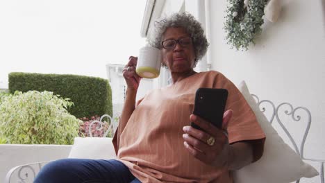 Senior-african-american-woman-using-smartphone-while-drinking-coffee-sitting-on-the-porch-of-the-hou