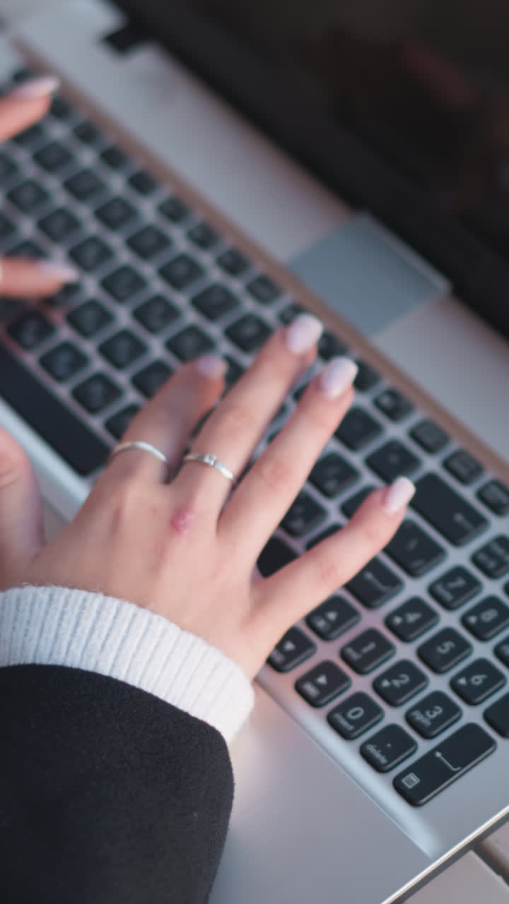 primer plano de manos escribiendo en el teclado de la computadora portátil, adornado con anillos de plata y clavos pulidos, cómodas mangas de suéter visibles, con el teléfono inteligente colocado cerca de la laptop en una mesa de madera