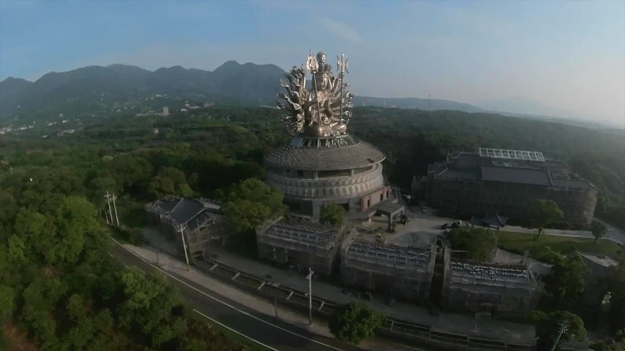 Aerial orbiting shot of Massive Guanyin goddess silver statue in China with many arms and two scepters. Blue sky and mountain range in background.