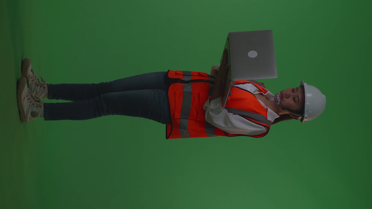vista lateral de todo el cuerpo ingeniera asiática con casco de seguridad trabajando en una computadora portátil mientras está de pie en el estudio de fondo de pantalla verde, durante la puesta o salida del sol