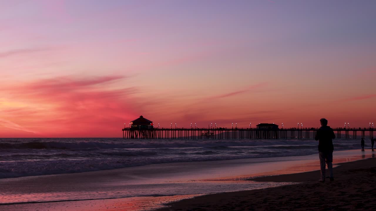 한 남자는 미국 캘리포니아주 서핑 도시의 헌팅턴 비치 부두를 배경으로 화려한 빨간색, 보라색, 감귤, 분홍색, 파란색 일몰을 배경으로 해변에서 휴가를 즐깁니다.