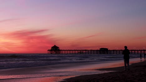 Un-Hombre-Disfruta-De-Sus-Vacaciones-En-La-Playa-Durante-Una-Hermosa-Puesta-De-Sol-Roja,-Púrpura,-Mandarina,-Rosa-Y-Azul-Con-El-Muelle-De-Huntington-Beach-Al-Fondo-En-Surf-City-USA-California