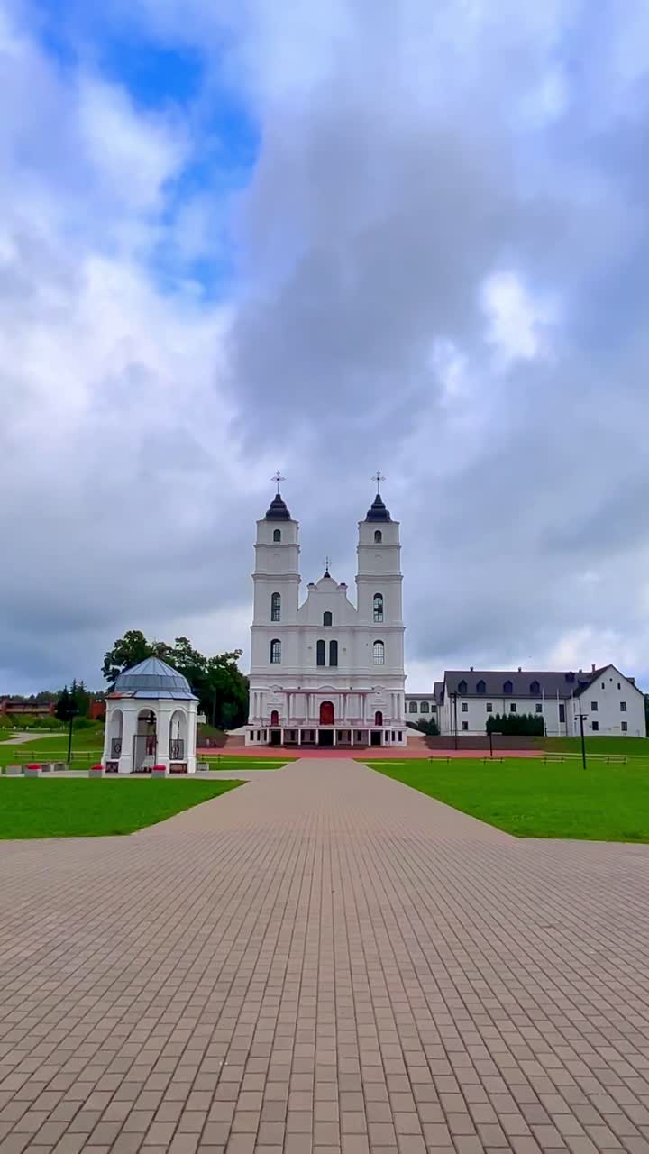 Maestosa Basilica di Aglona in Lettonia che si erge imponente contro un cielo nuvoloso. Le sue due torri bianche gemelle e la grande facciata sono centrali, con un sentiero lastricato che conduce all'ingresso e prati verdi su entrambi i lati.