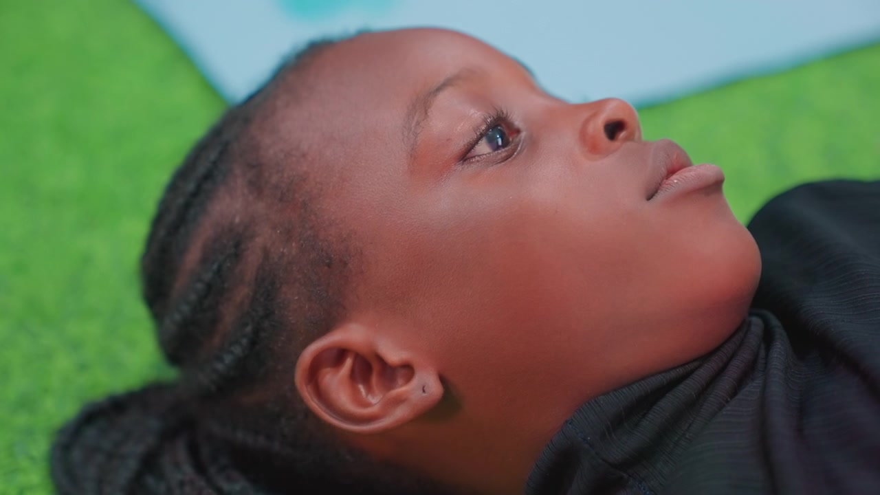 Black child lying on grass thoughtful, sideprofile closeup with pensive gaze, quiet mood, braided hair, soft green backdrop, contemplative student daydreaming about future, calm reflective atmosphere