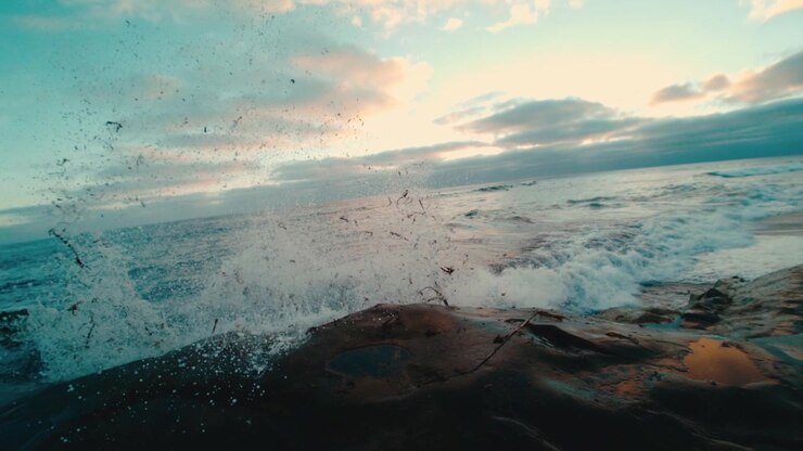 Immersive low-angle shot of waves surging over a rocky shoreline with droplets speckling the lens at golden sunset, coastal nature seascape travel themes.