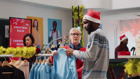 african american worker wearing santa hat helping woman browsing for clothes in festive christmas decorated clothing store. employee assisting elderly customer in festive adorn fashion shop