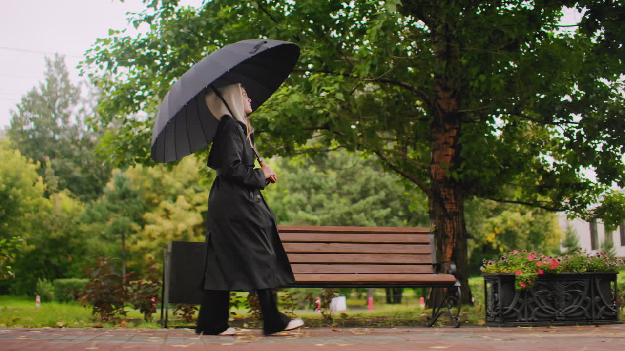 Young woman in long black coat and hood walking with umbrella through park on rainy day, surrounded by green trees and wet pavement, peaceful autumn lifestyle scene in natural outdoor environment
