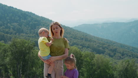 tired woman with little children stand against green hills