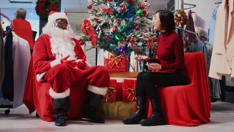 Afroamerikanischer-Angestellter,-Der-Als-Weihnachtsmann-Fungiert-Und-Sich-Neben-Dem-Weihnachtsbaum-Mit-Einer-Asiatischen-Frau-In-Einem-Weihnachtsgeschäft-Für-Verzierte-Kleidung-Setzt.-Modegeschäftsmitarbeiter-Bietet-Dem-Kunden-Ein-Geschenk-An