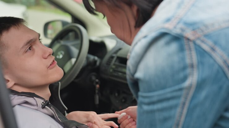 Adolescente bianco curato all'interno dell'auto, madre in denim appoggiata al veicolo, che porge un oggetto e gli sostiene la bocca, interazione di assistenza intima, interni dei sedili e ambiente stradale, famiglia affettuosa