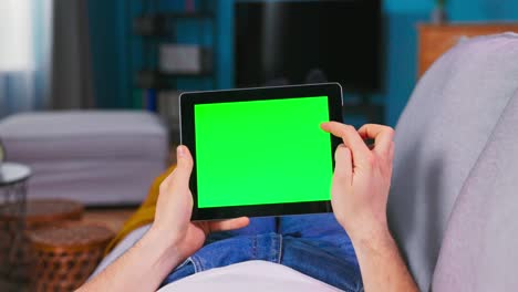 hombre joven en casa descansando en un sofá usando con pantalla de maqueta verde tableta computadora en modo paisaje horizontal. hombre usando gestos con dispositivo de pantalla táctil, navegando por internet, viendo contenido