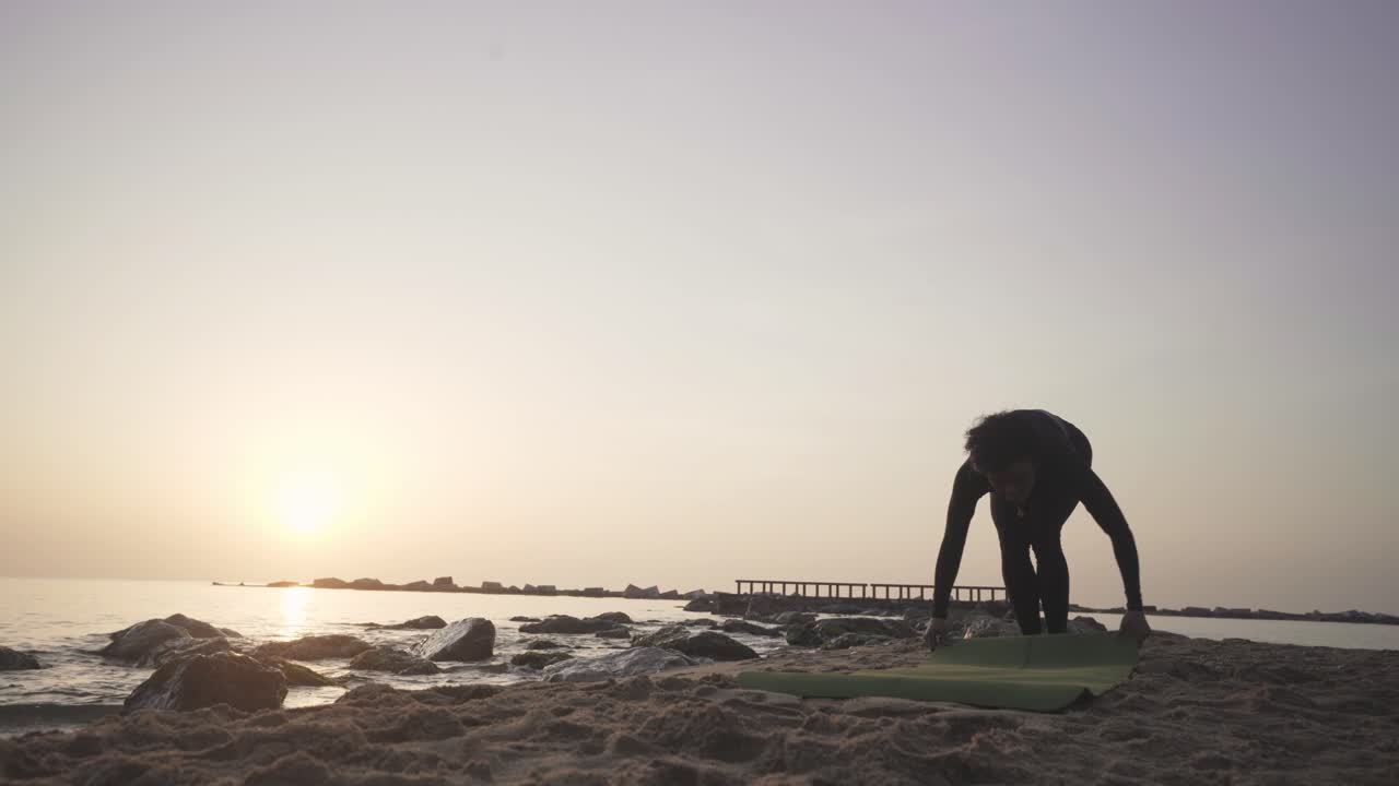 젊은 여성은 일출 때 아침 운동을 시작할 준비가 된 모래 해변에서 요가 매트를 펼칩니다.