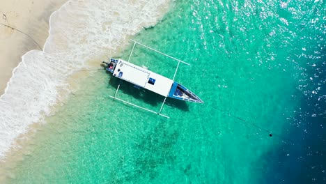 Un-Pequeño-Bote-Sentado-En-La-Superficie-De-Aguas-Azules-Brillantes,-Anclado-A-La-Orilla-De-Arena-Blanca-De-Una-Playa-Tropical-En-Indonesia,-Vista-Superior