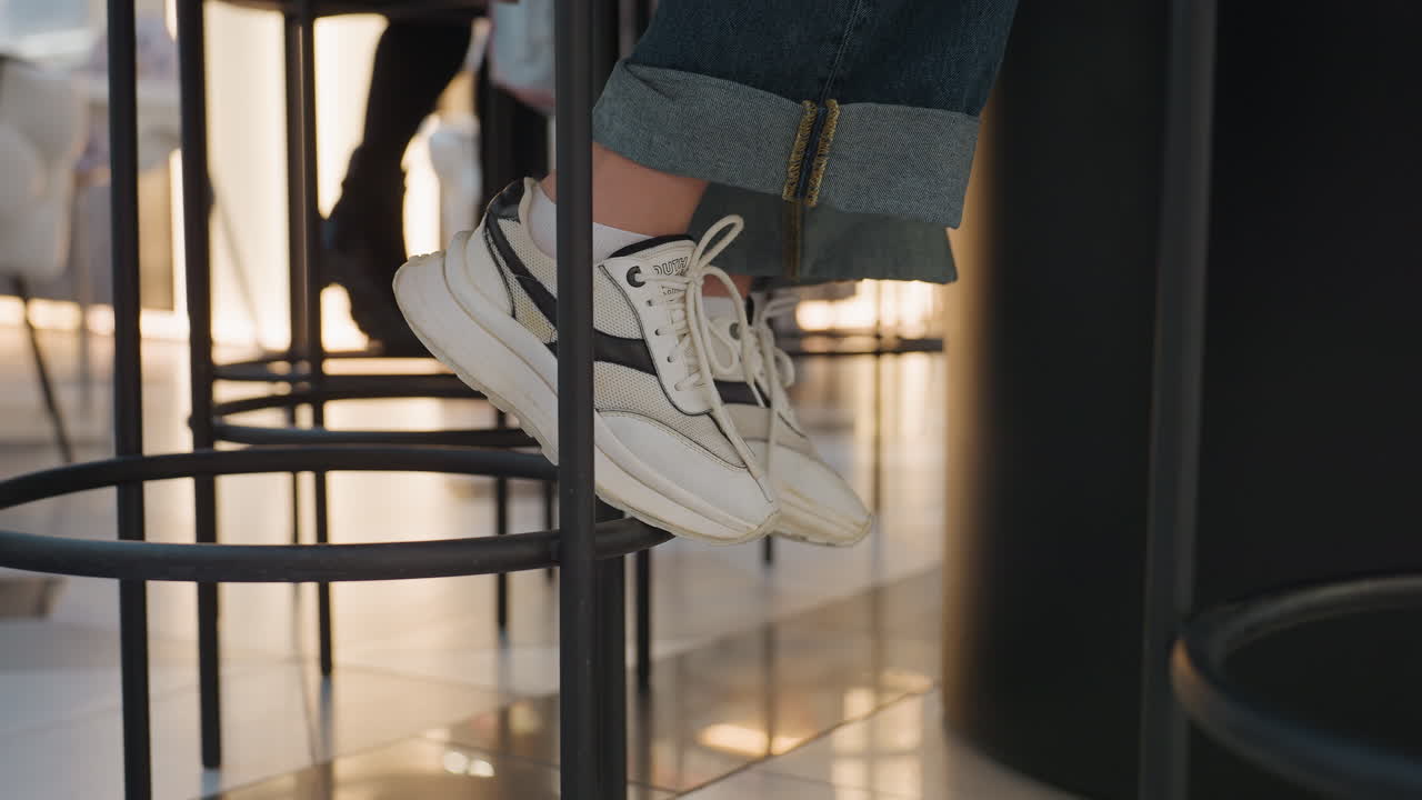 높은 테이블에 흰색 스니커즈가 매달려 있고, 동료들이 메모와 커피를 나누며, 금속 링에 미묘하게 발을 움직이고, 윤기 있는 바닥에 반사되며, 편안한 저녁 대화와 편안한 바디랭귀지, 따뜻함