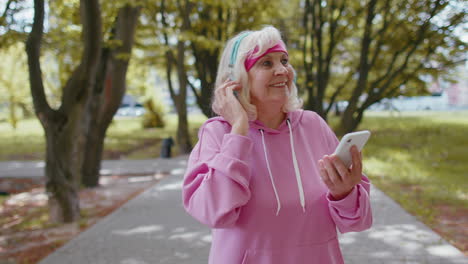 Athletic-senior-sport-runner-woman-wearing-earphones-listening-music-from-smartphone-in-morning-park