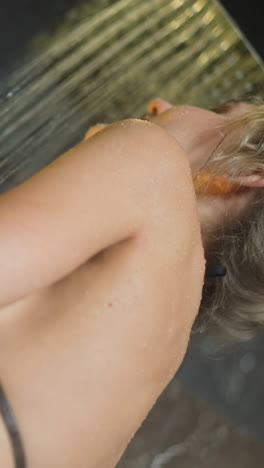 young woman with fair hair bun stands under running water washing slim body in contemporary shower booth at early morning daily routine slow motion