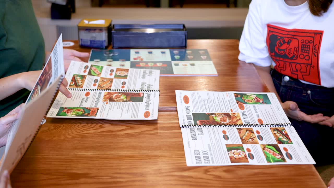 木製のレストランのテーブルに座り、メニューの品を指差している食事客の真上からの眺め。