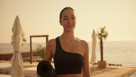 Portrait-of-a-brunette-girl-in-a-black-top-who-holds-a-black-yoga-mat-in-her-hands,-looks-at-the-camera-and-smiles-against-the-backdrop-of-a-beach-with-umbrellas-and-palm-trees