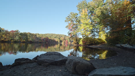 A-rising-shot-of-a-calm-lake-surrounded-by-autumn-trees-with-large-stones-and-a-small-beach-in-the-foreground-in-this-New-England-scene