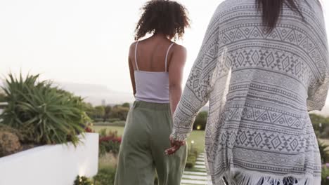 rear view of happy biracial lesbian couple holding hands and walking in garden, slow motion