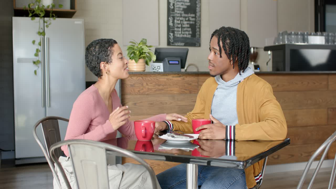 Diverse couple talking eagerly nodding cradling red mugs and reassuring partner at table in kitchen