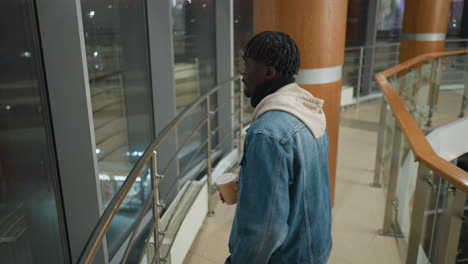 back view of young man walking through modern indoor balcony walkway with reflective windows, wooden columns, and stainless railing at night, dressed in hoodie and denim jacket, calm atmosphere