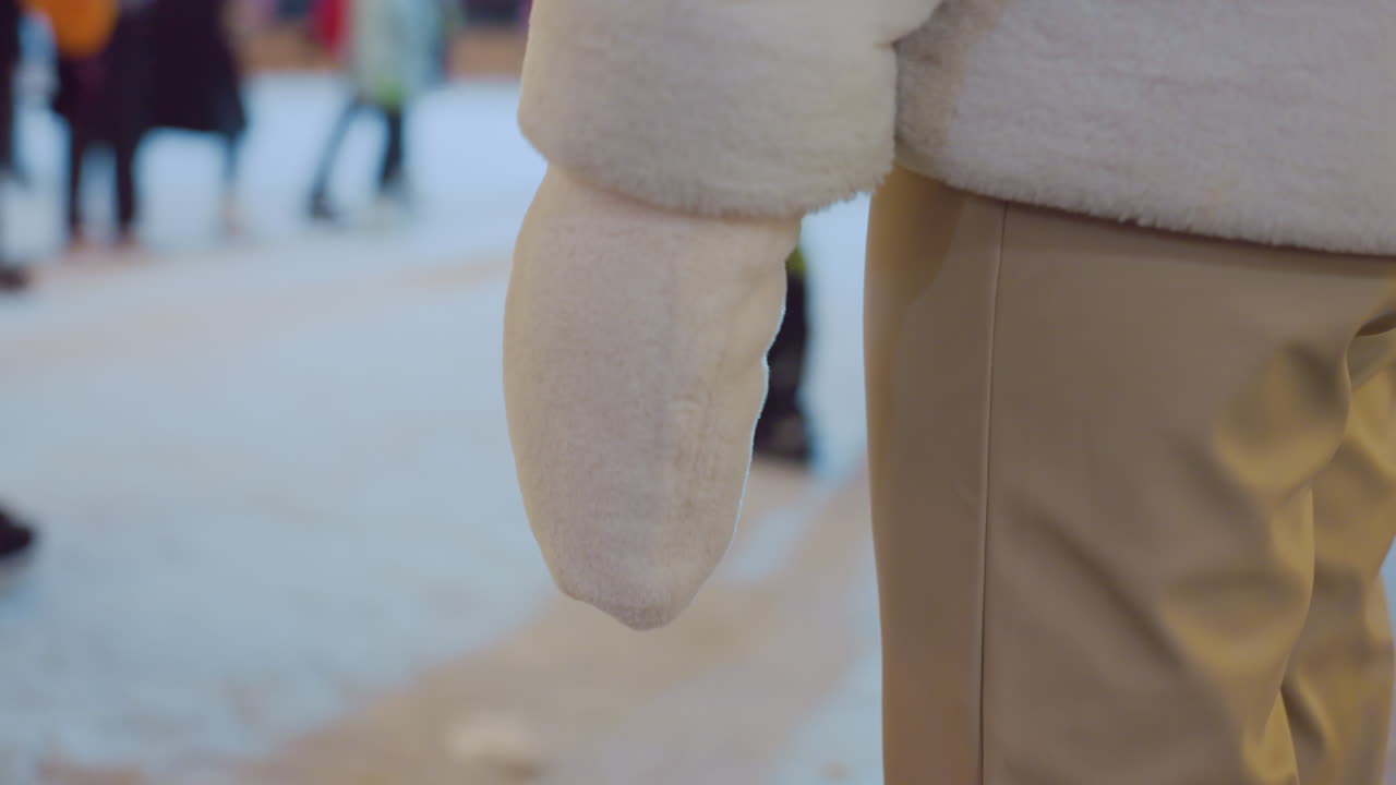 Rear view of person in brown trousers and winter coat standing still before gently turning around as people skate and move in the background during winter