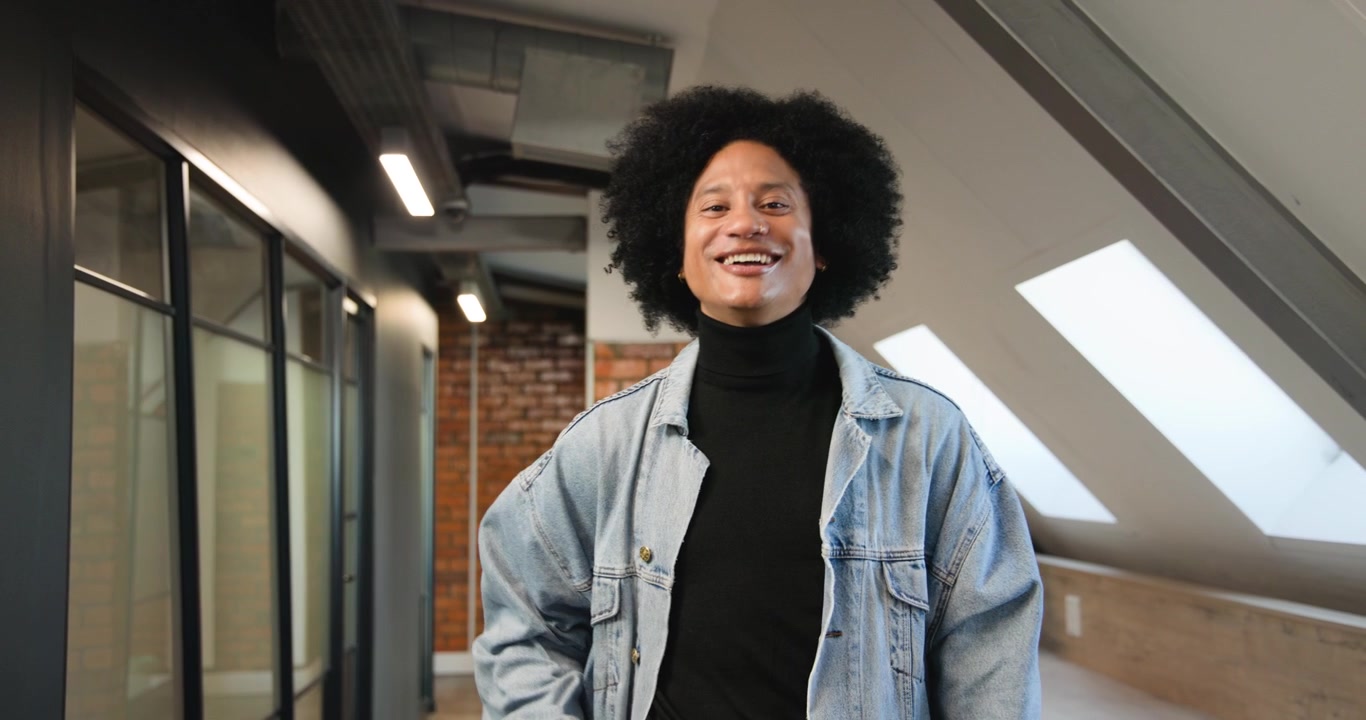 Uomo afroamericano in denim che nota il lucernario del loft, osserva la scrivania, si mette comodo e sorride alla telecamera.