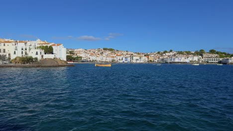 Cadaqués-Ciudad-Mediterránea-En-La-Costa-Brava-De-Girona,-Vista-Del-Intenso-Mar-Azul-Y-La-Línea-De-Casas-Blancas,-Y-Un-Barco-Amarillo-En-El-Agua