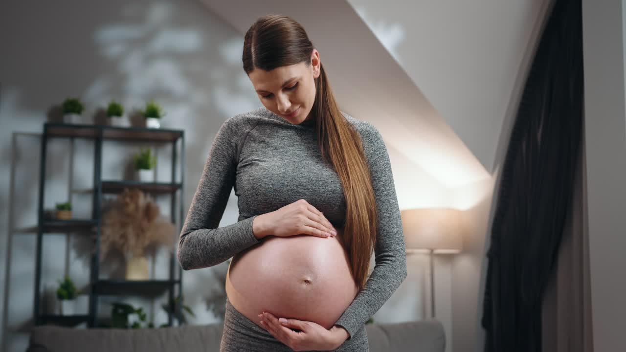 Young woman cradles her pregnant belly with gentle hands looking down with a soft expression in a