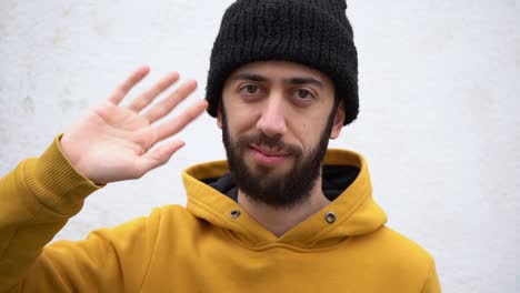 Caucasian-Man-In-His-20s,-Wearing-Black-Beanie-Hat-And-Yellow-Jacket,-Hand-Waving-Hello-To-Camera
