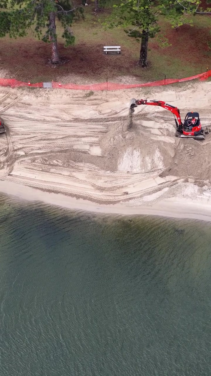 Vista aerea di macchinari pesanti che spostano sabbia durante la riqualificazione di una spiaggia lungo un lungocanale residenziale.