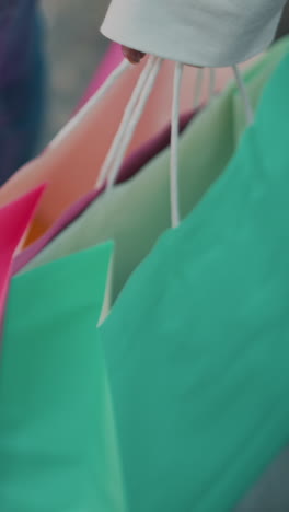 primer plano de una persona sosteniendo bolsas de compras verdes y rosas mientras está de pie en el suelo de mármol brillante, zapatillas blancas borrosas en el fondo mejoran el ambiente minorista moderno con el movimiento sutil de las bolsas oscilantes