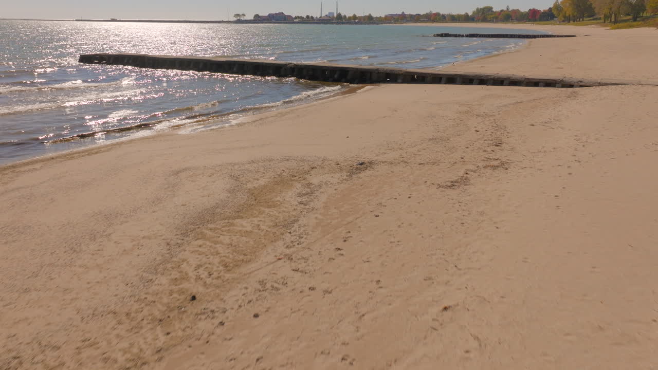 Drone aerial flying low from the beach over a concrete pier and Lake Michigan in Sheboygan, Wisconsin, as sunlight shimmers on gentle autumn waves on a clear, beautiful day