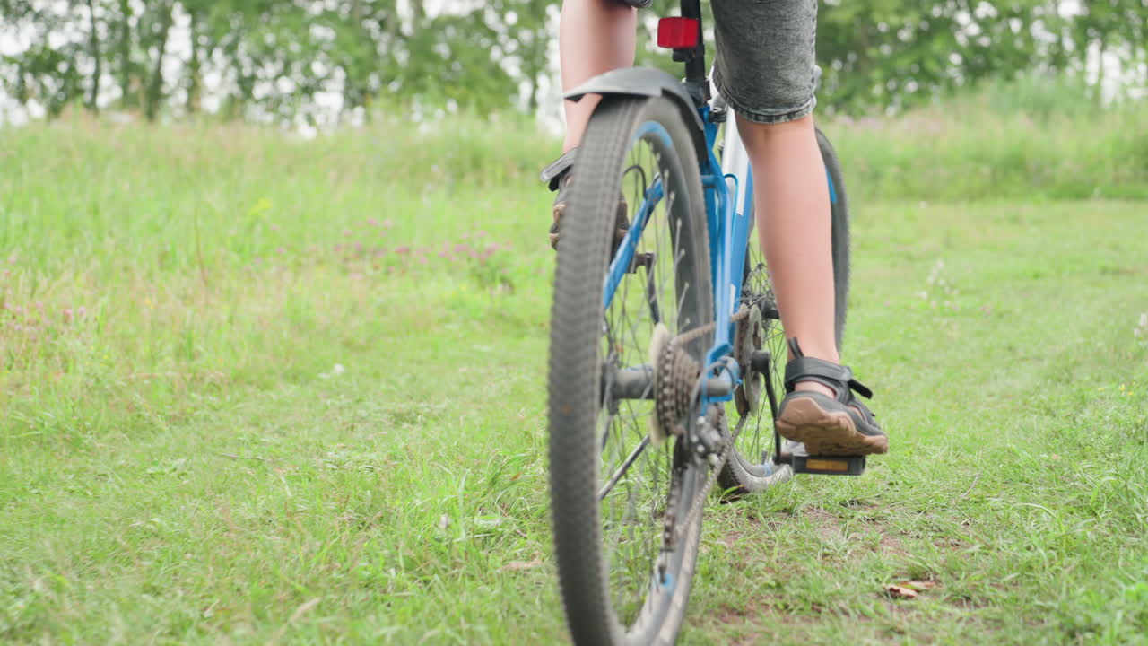 코카서스 아이가 풀이 무성한 길에서 파란색 자전거를 타고 멀어져 가고, 페달이 돌아가고 뒷바퀴가 여름 들판을 가로지르며 회전하고, 진흙 묻은 신발과 움직이는 체인이 보이고, 넓은 녹색 풍경과 수평선이 펼쳐져요.