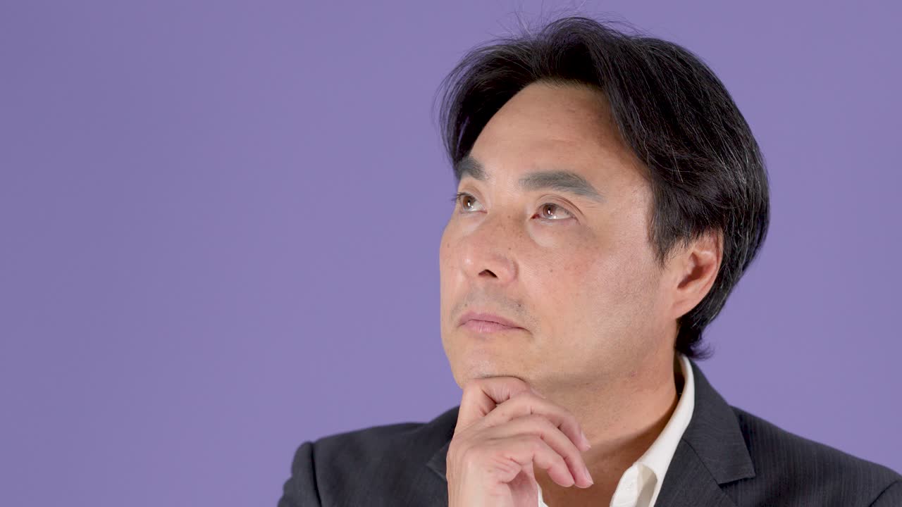 スーツを着た中年のビジネスマンが、顎に手を当て、わずかに頭を動かしながら、思慮深くポーズを取る。スタジオ照明。