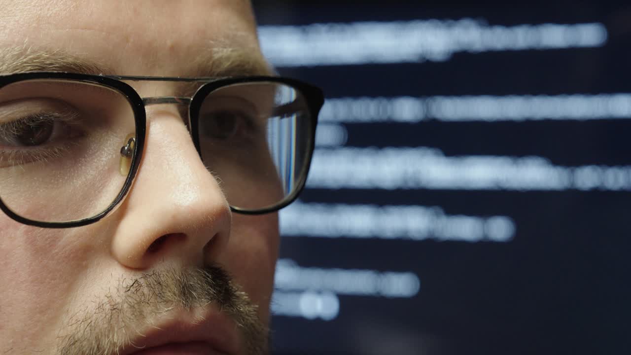 A man with glasses in close-up, focused, with a large display behind him showing AI programming code