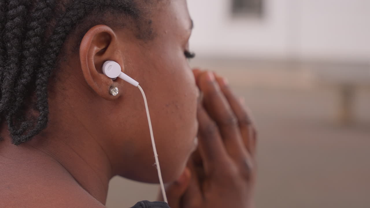 Corritrice concentrata che ascolta musica all'aperto. Sportiva urbana che si prepara con calma con le cuffie vicino al viso. Atleta con trecce che si concentra pacificamente prima dell'esercizio in un ambiente urbano all'aperto.