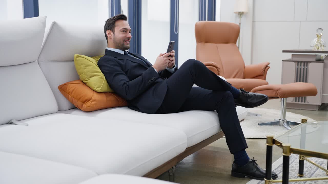 Calm and happy businessman chatting on phone enjoying a moment of leisure or satisfaction due to receiving good news or feeling accomplished lounging on white sofa with crossed legs at premium office