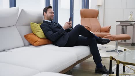 calm and happy businessman chatting on phone enjoying a moment of leisure or satisfaction due to receiving good news or feeling accomplished lounging on white sofa with crossed legs at premium office